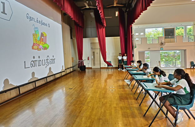 Students in uniforms sit at desks in a school hall facing a projected Tamil language slide with colorful text.